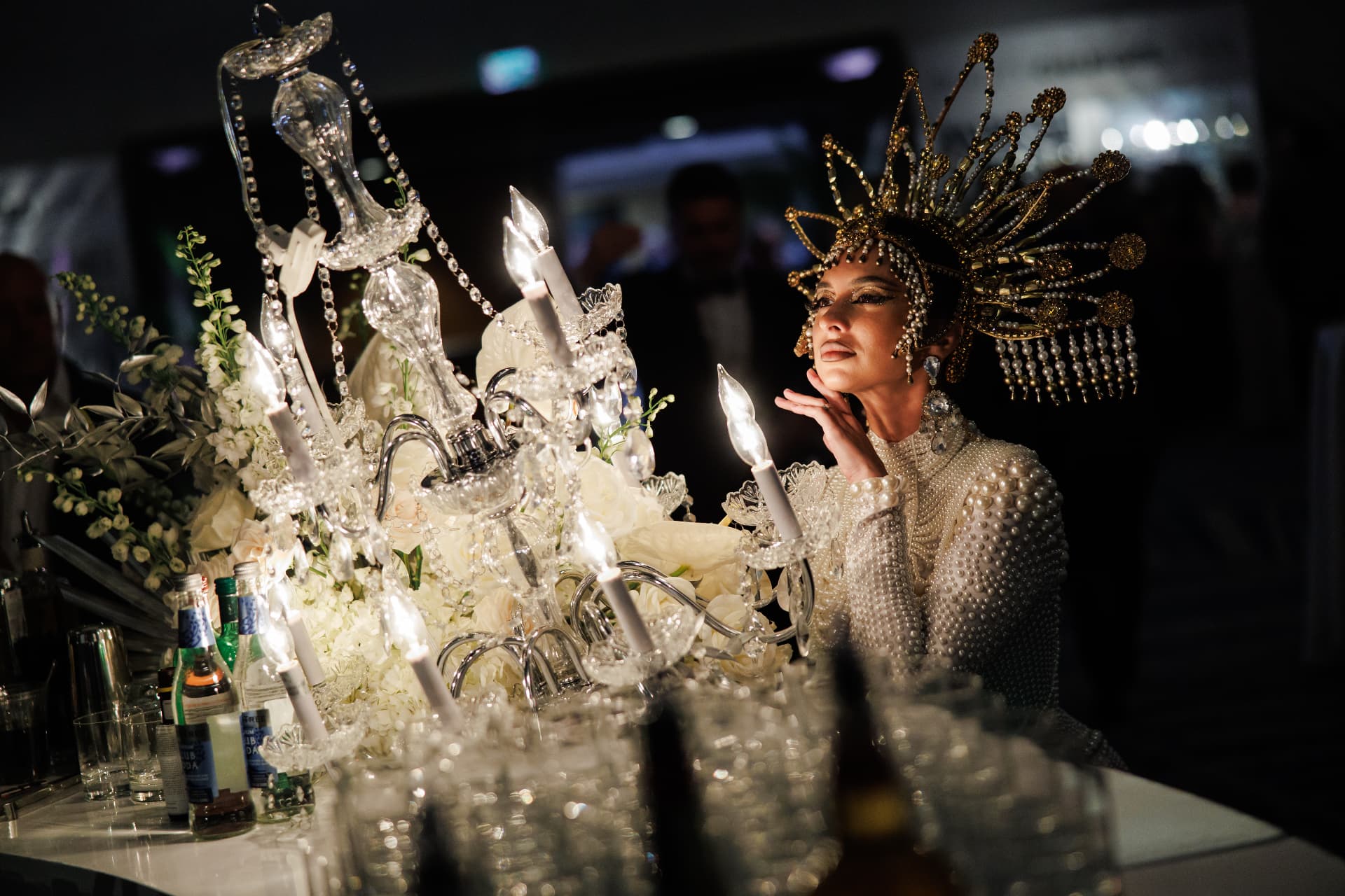 Performer in an elaborate crystal headpiece beside a glowing chandelier at a luxury event
