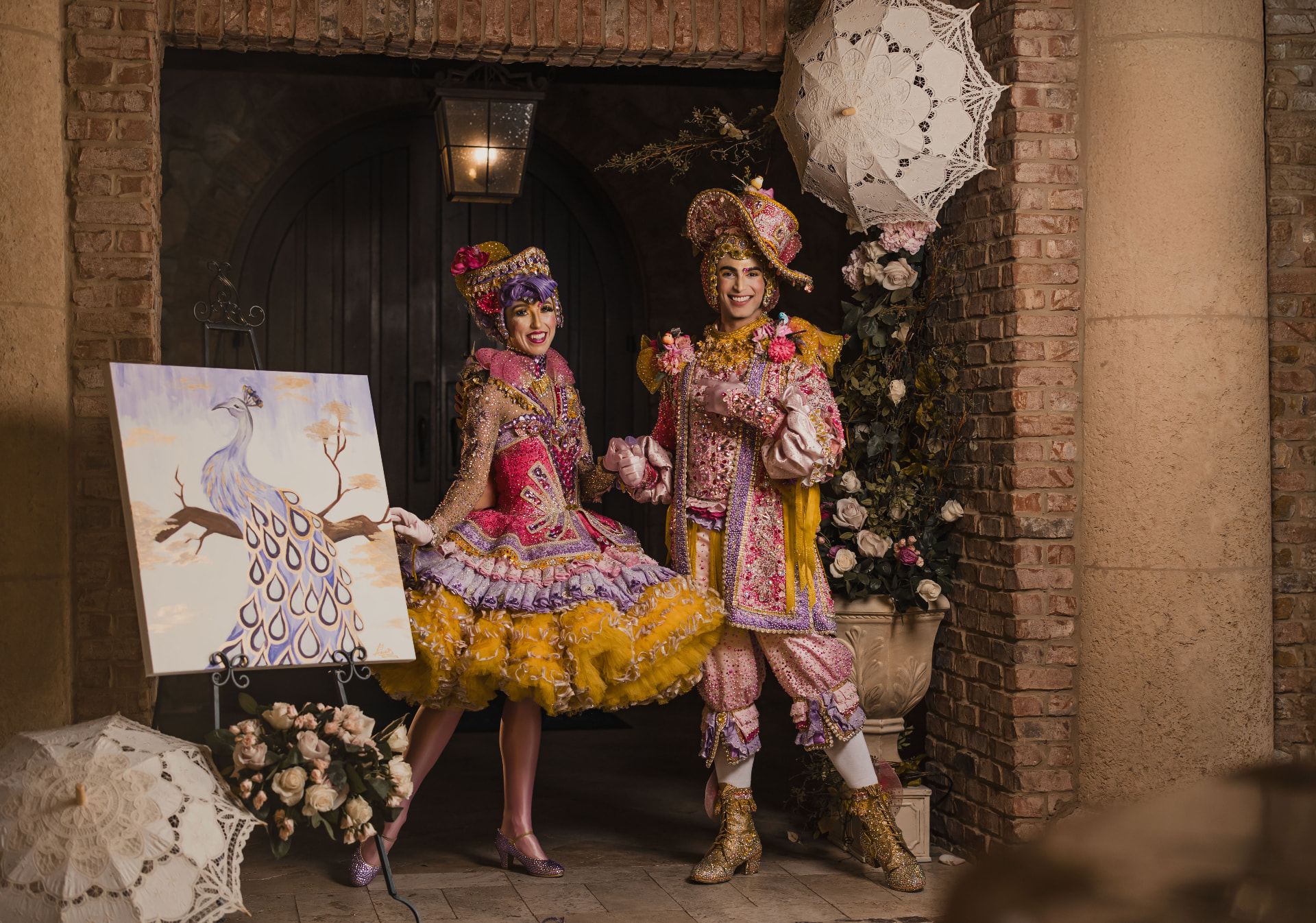 Two costumed arrival performers in ornate jewel-toned couture beside a painted peacock canvas